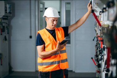 Adult electrician builder engineer testing and screwing equipment in fuse box and repairing of modern electricity power station using data from the tablet.. Automatic control cabinet clipart