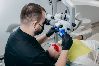 Erkek dişçi modern mikroskopla hastanın dişlerini inceliyor.