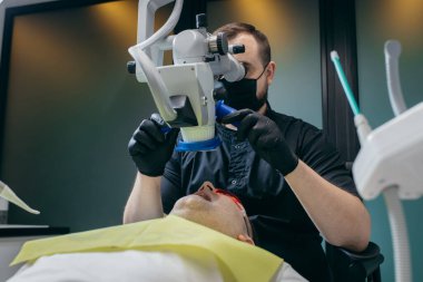 Erkek dişçi modern mikroskopla hastanın dişlerini inceliyor.