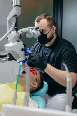 Erkek dişçi modern mikroskopla hastanın dişlerini inceliyor.