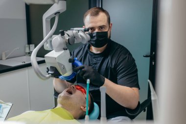 Erkek dişçi modern mikroskopla hastanın dişlerini inceliyor.