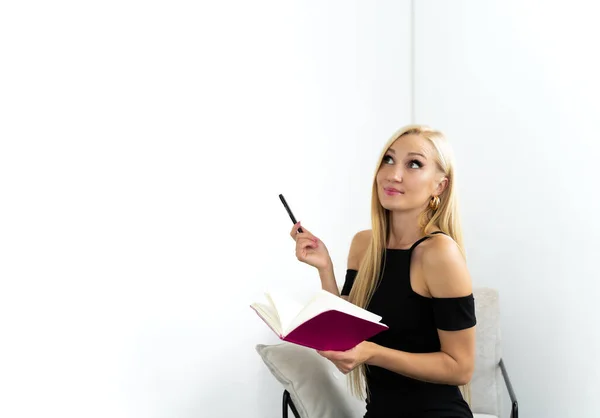 Image of young european woman, company worker, smiling and holding pen and notepad while sitting against white background