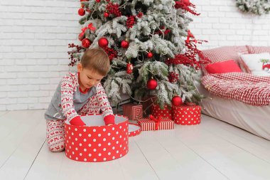 Süslü bir Noel ağacının yanında yeni yıl pijaması giymiş bir çocuk ve kırmızı renkli bir yatak hediye paketini açıyor. Çocuk yeni yıl için bir hediye tarafından şaşırtılıyor. duygular