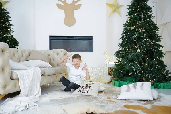 one European young boy in casual clothes fair-haired with brown eyes sits in a lotus position happy on Christmas Eve in a room near the Christmas tree by the fireplace with beautiful pastel pillows