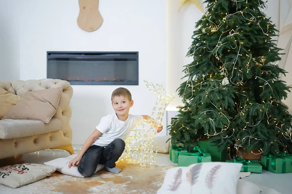 one european young boy in casual clothes, fair-haired with brown eyes, hugs a christmas decoration of a luminous deer in a new year\'s room near the christmas tree by the fireplace with beautiful paste