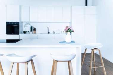 Stylish interior design of a modern fashionable snow-white kitchen in a minimalist style with an island and two bar stools, huge floor-to-ceiling windows and a glass counter