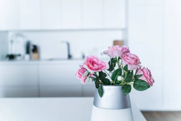 In a white kitchen, pink rose flowers in a vase in the foreground stand on a desktop. Concept interior, space, inspiration, close-up, space for text