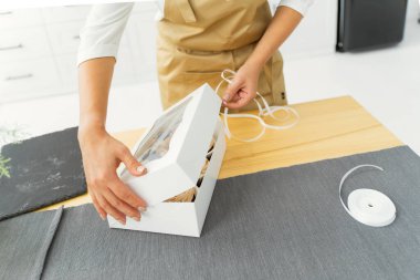 A pastry chef in a white kitchen in a beige apron closes a box of sweets. Small business concept, confectioner, cook, holidays, birthday, Christmas, Easter, New Year