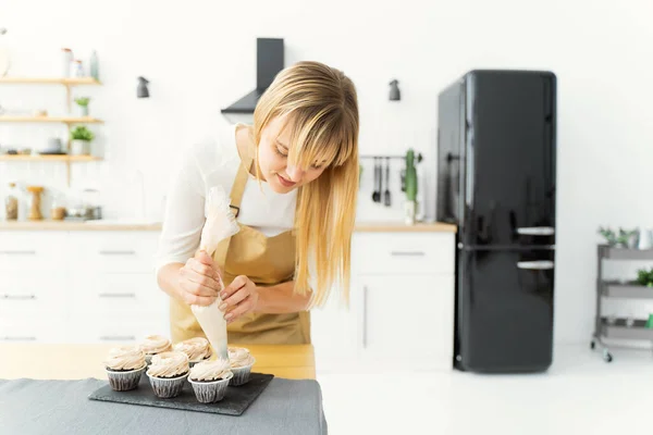 Female pastry chef in beige apron with chocolate cupcakes, decorating with cream cheese frosting, in her kitchen at home indoors. Women\'s business, concept