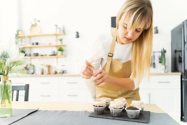 A pastry chef in a beige apron is cooking with chocolate cupcakes decorated with cream cheese frosting in her kitchen at home indoors. Women's business, concept