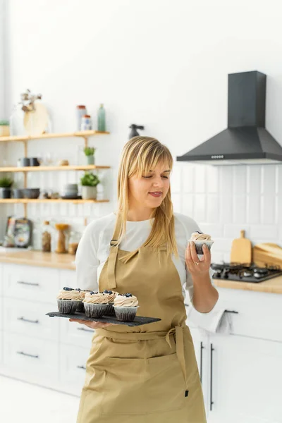 A European girl confectioner stands in a bright kitchen with green flowerpots and holds a tray of cupcakes in one hand, and holds one cake in the other and stares at it with a smile
