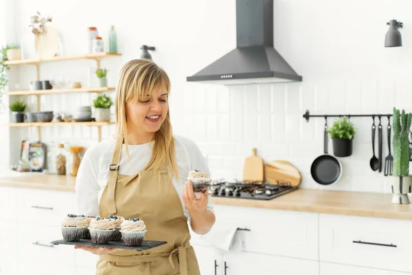 A European girl confectioner stands in a bright kitchen with green flowerpots and holds a tray of cupcakes in one hand, and holds one cake in the other and stares at it with a smile