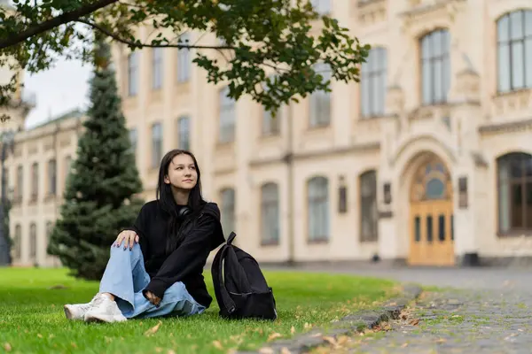 Bir kız öğrenci üniversitenin yanındaki çimlerde oturur. Yüksek kalite fotoğraf