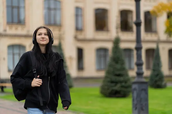 stock image A girl student goes to campus with a backpack on her shoulders and listens to music in headphones. High quality photo