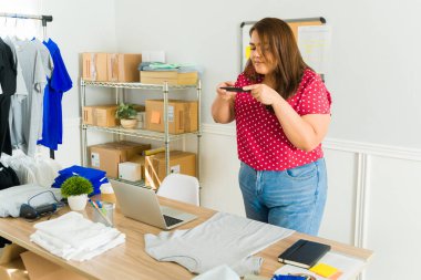 Çevrimiçi dükkanı ve sosyal medya için akıllı telefonuyla tişört satan genç bir kadın. 