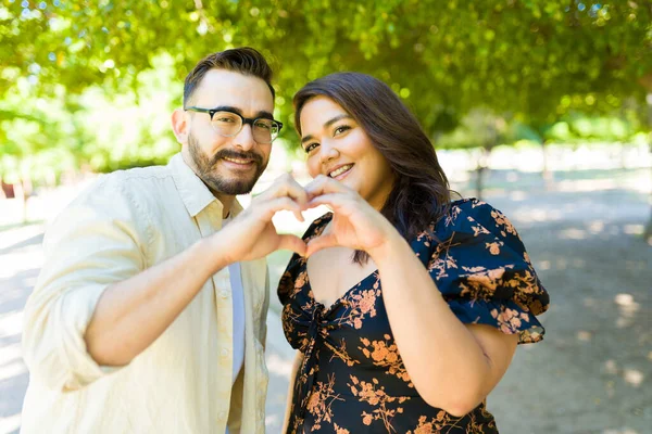 Happy smiling couple in love making a heart gesture with their hands outdoors and looking at the camera