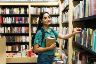 Kitapçıda sırt çantalı mutlu bir İspanyol genç kadın akademik kitaplar alırken göz teması kuruyor. 