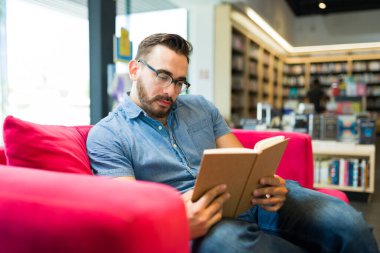 Yakışıklı beyaz adam kırmızı bir koltukta oturuyor ve kitapçıda kitap okuyor.
