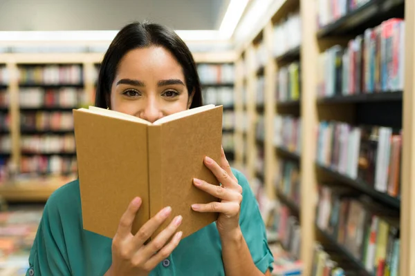 在书店看小说时 快乐的年轻女人躲在书本后面笑着 — 图库照片