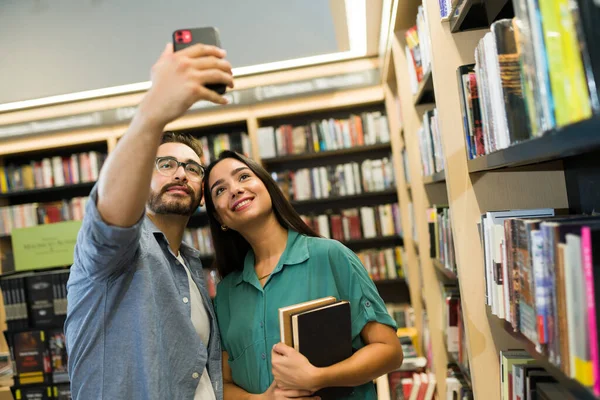 Attractive happy couple taking a selfie together with a smartphone while buying books at the bookstore