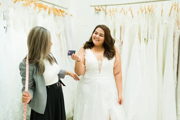 Happy obese young woman buying her beautiful wedding dress with a credit card and talking to a saleswoman at the bridal shop