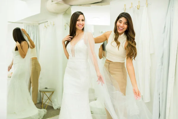 Cheerful future bride with a wedding gown and veil laughing with a friend while trying on dresses at the bridal shop