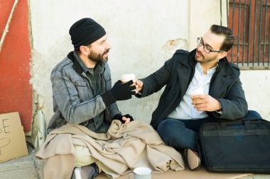 Çekici bir genç adam, sakallı evsiz bir adamla kahve ve yemek paylaşırken aynı zamanda nezaket sergiliyor.