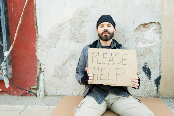 Portrait of a miserable homeless man living on the streets showing a cardboard sing asking for help for food or money