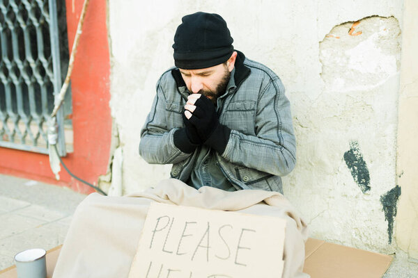 Cold homeless man warming his hands wearing gloves and holding a sign of please help 