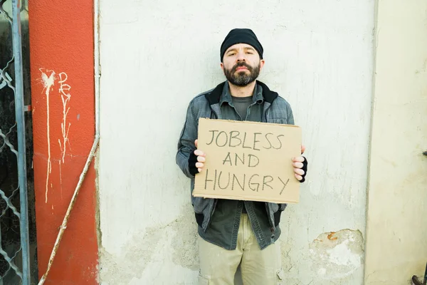 貧困の中で暮らすヒスパニック系のホームレスの男が助けを得るために失業と空腹の段ボールのサインを示しています — ストック写真