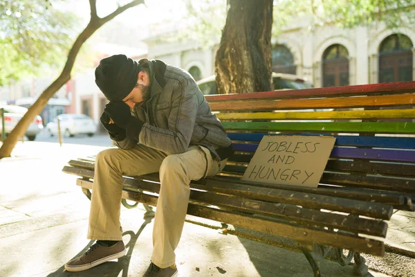 忧心忡忡的无家可归的人坐在公园的长椅上 上面有一个失业和饥饿的纸板标志 — 图库照片