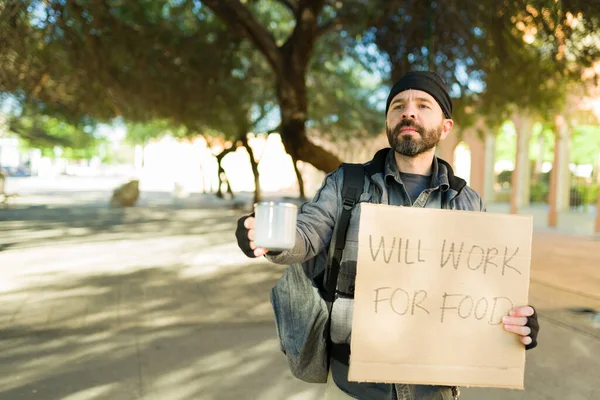 Poor homeless man begging for money with a cup while holding a sign will work for food
