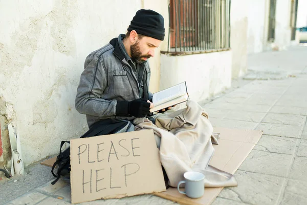 在街上挣扎的时候 一个留着胡子的无家可归的男人一边看书 一边展示着一张请帮他签牌的纸牌 — 图库照片