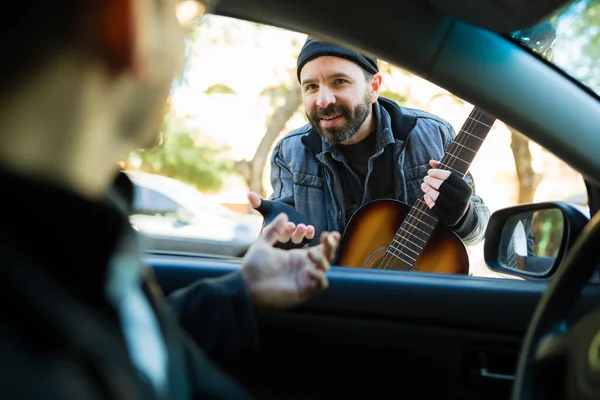 路上でギターと音楽を演奏しながらドライバーからお金を求めながら 髭を生やしたホームレスの男を笑顔 — ストック写真