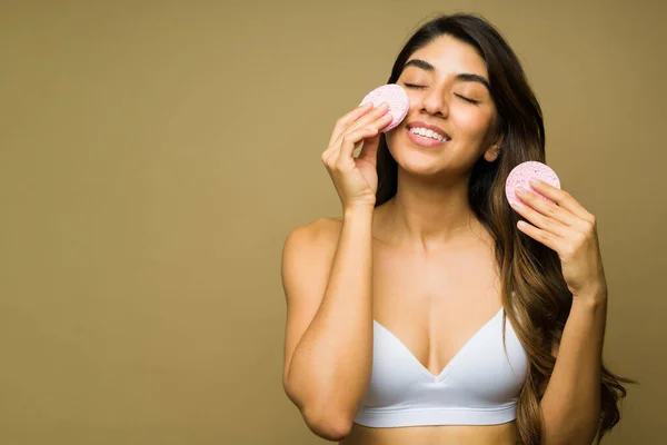 Attractive relaxed latin woman smiling while doing a skin exfoliation with beauty sponges and skincare products