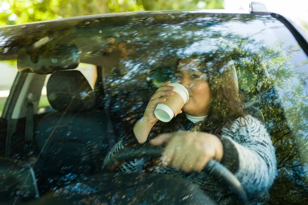 Happy fat hispanic woman seen through the windshield drinking coffee and driving the car