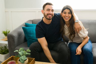 Çekici genç çift evdeki kanepede dinlenirken sarılıp gülümsüyor ve göz teması kuruyor.