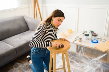 Güzel bir kadın tabure mobilyalarında zımpara kağıdı kullanıyor ve bir ev geliştirme projesi üzerinde çalışıyor.