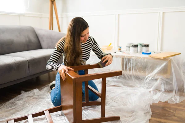 古代の椅子の上にサンドペーパーを使用してヒスパニック系の若い女性が彼女の改装事業のために家でめくる家具をやっている — ストック写真