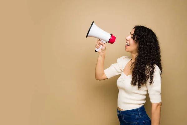 Profile of an excited young woman with curly hair using a megaphone and shouting a message next to copy space ad