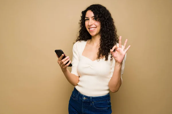 Happy hispanic woman with curly hair doing a perfect ok sign while using her smartphone texting