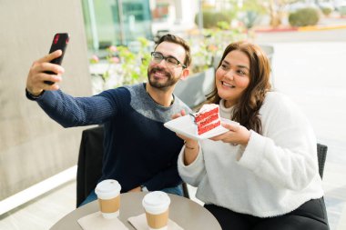 Attractive influencer fat woman and smiling man taking a selfie for their social media content while eating cake and drinking coffee 