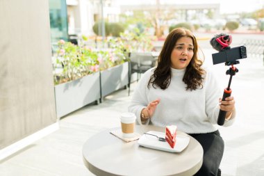 Mexican chubby woman and content creator talking to the camera while filming a video while drinking coffee and eating dessert