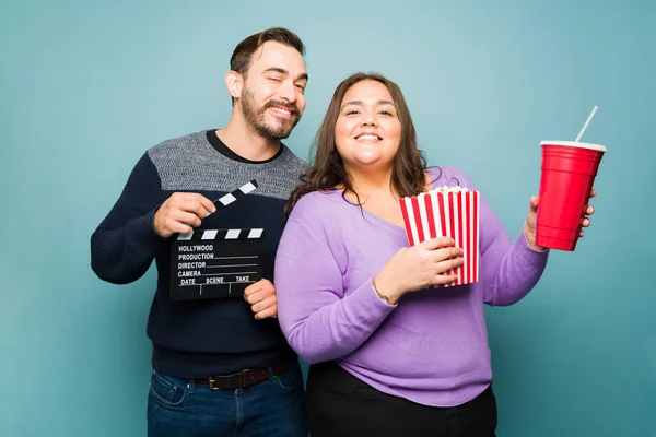 Happy beautiful couple loving the movies while eating popcorn and using a clapperboard against a blue background