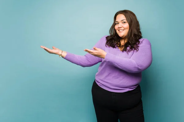 Cheerful latin obese woman smiling while showing a product or ad against a blue studio background