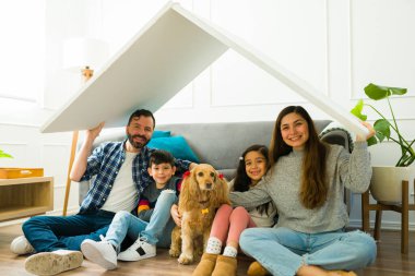 Excited beautiful family with children and home owners holding a roof above their heads after buying or moving to their new house with a beautiful dog