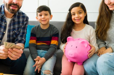 Beautiful young boy and little girl smiling with their parents while saving their allowance money in the piggy bank