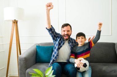 Cheerful dad and happy little boy cheering and celebrating while watching the soccer game together on tv