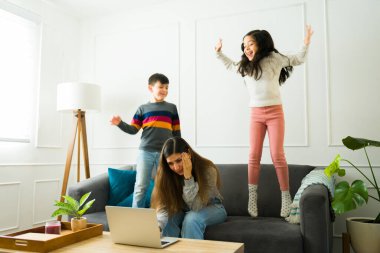 Stressed young mom trying to work on the laptop at home while taking care of her young kids jumping and playing on the sofa 