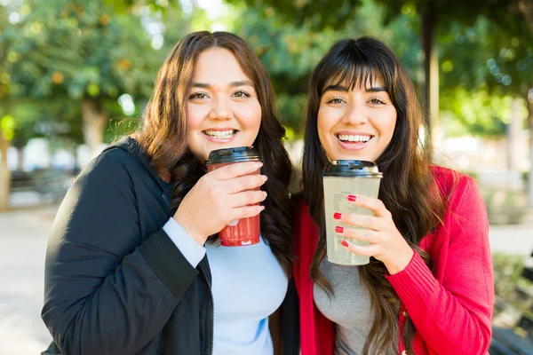Excited latin women friends laughing and having fun while drinking coffee hanging out at the park